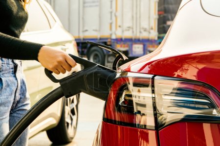 Junge EV- oder Elektroauto-Fahrerin hält Gleichstromstecker an ihrer roten Elektroauto-Batteriesteckdose in der Tankstelle: Sauberes Energiefahrzeug Alternative Energie wird beliebtes Konzept.