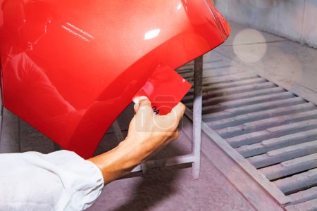 Photo for Hand of a male auto painter working car painting room inspects and compares the red paint to the sheet metal sprayed front bumper to get similar and beautiful color : Jobs in auto repair garages. - Royalty Free Image