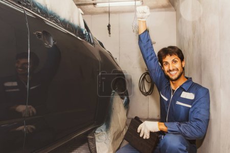 Photo for Handsome skilled car painter dressed in automotive painter's uniform sits happily holds up his fist admires the new blue paint masterpiece in the paint room smiling and looking into the camera. - Royalty Free Image