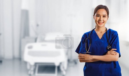 Portrait healthy asian female surgeon in blue dress with stethoscope standing crossed hands smiling friendly looking at camera all standing smartly in abstract medical facility patient room.