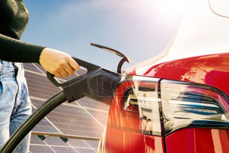 Mano de mujer joven conduciendo un vehículo eléctrico del EV que sostiene el cable del enchufe de DC al enchufe rojo de la batería del vehículo eléctrico que carga la electricidad de la fuente de energía solar de la célula solar.