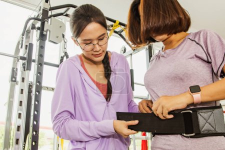Foto de Las mujeres entrenadoras aconsejan y apoyan el ejercicio Las mujeres mantienen su salud ayudándolas a usar cinturones de levantamiento de pesas para prevenir el dolor más bajo y lesiones abdominales cuando se inclinan hacia abajo. - Imagen libre de derechos