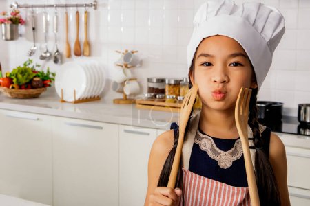 Photo for Happy portrait asian girl cute in white chef hat and apron preparing to cook in the kitchen playful innocently holding cooking utensils by her cheek, smiling and looking at the camera in a good mood. - Royalty Free Image