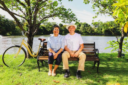 Foto de Senior asiático jubilado pareja saludable y alegre viaje salud parque estacionamiento bicicleta sentado banco en un jardín al aire libre claro tiempo siguiente sombrío estanque mirando a la belleza dentro del increíble jardín. - Imagen libre de derechos