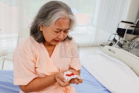 Foto de Ancianos mujer asiática estresada, llorar preocupación sentado en una cama hospital sala de emergencias que sufren de neumonía tos que la puso en riesgo de cáncer de pulmón se sorprendió al ver la sangre en su esputo. - Imagen libre de derechos