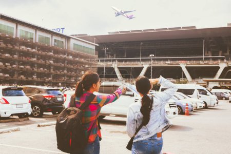 Two asian women wheeling suitcases and backpacks walked out of an airport parking lot to board passenger plane pointing departure terminal make appointment and check in their travel tickets.
