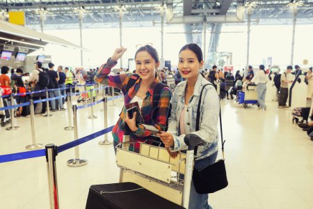 Two beautiful asian women in the counter overseas check in departure terminal preparing their suitcases board the plane holding their passports and plane tickets standing and looking at them.
