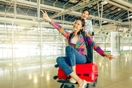 Lustig am Flughafen, während man im Flugzeug wartet, um ins Ausland zu reisen: Fröhliche asiatische Freundinnen sitzen auf Gepäckwagen mit ausgebreiteten Koffern und imitieren Vögel, die mit engen Freunden Spaß haben..