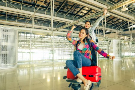 Heureux à l'aéroport en attendant de monter à bord de l'avion pour voyager à l'étranger : joyeuses amies asiatiques assises sur des chariots à bagages avec des valises étalées imitant les oiseaux qui s'amusent avec des amis proches.