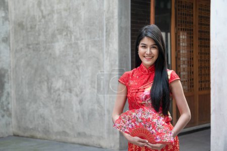 Foto de Retrato de imágenes de mujeres guapas en asia usando un cheongsam rojo sosteniendo un ventilador de madera en su mano con fondo de pared de hormigón, al concepto de chica china. - Imagen libre de derechos