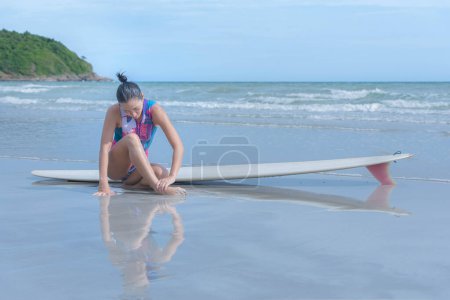 Asian woman sitting on the beach, she injured her ankle after exercising by surfing in the sea. with people's health care and the concept of surfboards