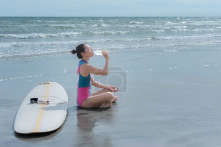 Junge Asiatin am Strand Sie trank Wasser aus einer Flasche, um ihren Durst zu stillen. Konzept des Surfbrettsports für die Gesundheit