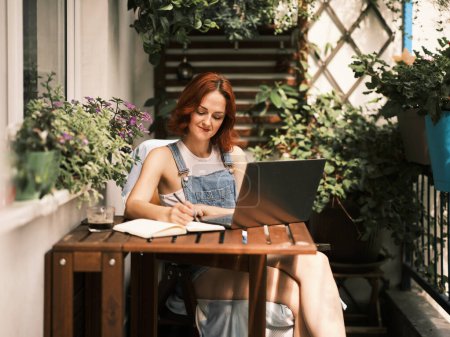 A red-haired woman focuses on writing in a notebook while sitting at a wooden table on a plant-filled balcony