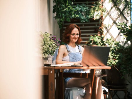 Téléchargez les photos : Une femme souriante en tenue denim travaille sur son ordinateur portable entouré de plantes sur un balcon ensoleillé - en image libre de droit