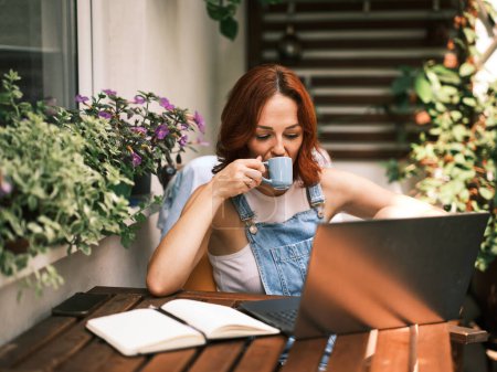 Téléchargez les photos : Femme rousse profite d'une tasse de café tout en utilisant son ordinateur portable sur un balcon ensoleillé avec des plantes luxuriantes - en image libre de droit