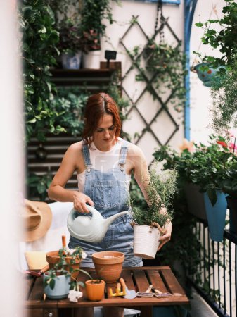 Photo for Woman in denim overalls lovingly watering her plants on a sunny balcony filled with greenery - Royalty Free Image