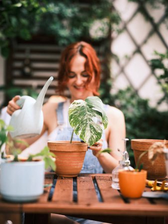 Téléchargez les photos : Femme souriante arrosant une plante feuillue distinctive sur un balcon équipé d'outils de jardinage et de pots - en image libre de droit