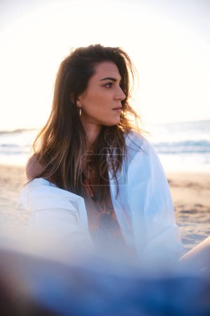 Téléchargez les photos : Une femme réfléchie s'assoit sur la plage, regardant loin contemplativement, avec le coucher de soleil en arrière-plan - en image libre de droit
