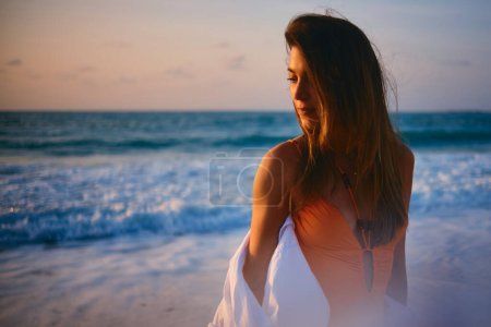 Photo for With her back to the camera, a woman in a dress looks at the ocean at sunset, symbolizing escape and longing - Royalty Free Image