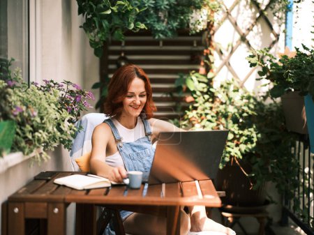 Téléchargez les photos : Une jeune femme aux cheveux roux aime travailler sur son ordinateur portable entouré de plantes en pot luxuriantes sur un balcon ensoleillé - en image libre de droit