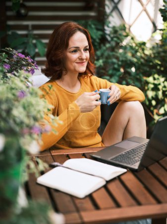 Warm gekleidete Frau genießt ihren Kaffee und ihren Laptop in einem lebendigen Garten zu Hause