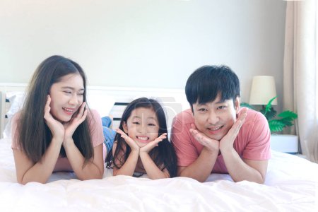 Photo for Happy Asian family has fun in bedroom. Father, mother and daughter girl lying on bed together, propping chin up with hands and smiling to camera, dad mom, parents having good memory with kid - Royalty Free Image