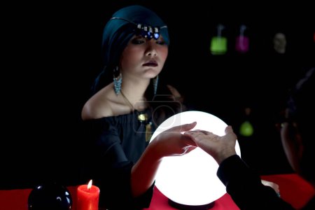 Photo for Mysterious magnificent beautiful woman fortune teller in black dress with magical crystal ball looking at customer palm, reading future, witch during fortune, palmistry and divination, magic ritual. - Royalty Free Image