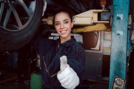 Beautiful female auto mechanic giving thumb up during checking wheel tires in garage, car service technician woman repairing customer car at automobile service, inspecting vehicle engine underbody.