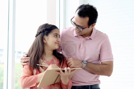 Photo for Cute Indian school student girl wears traditional dress near window, holds notebook pencil for doing homework, father teaching daughter kid at home, parent involvement in childhood education in family - Royalty Free Image