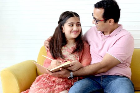 Photo for Cute Indian school student girl wears traditional dress sitting with father in living room and doing homework, dad teaching daughter kid at home, parent involvement in childhood education in family. - Royalty Free Image