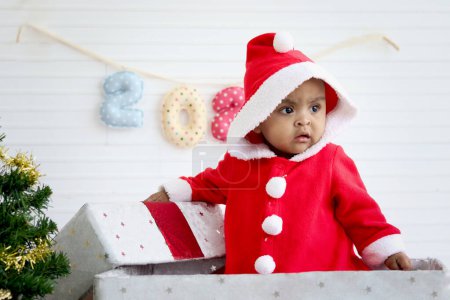 Photo for African baby kid in Santa Claus red costume standing in big gift box present at white living room, beautiful little child celebrates Christmas holiday. celebration, merry Christmas and happy new year. - Royalty Free Image