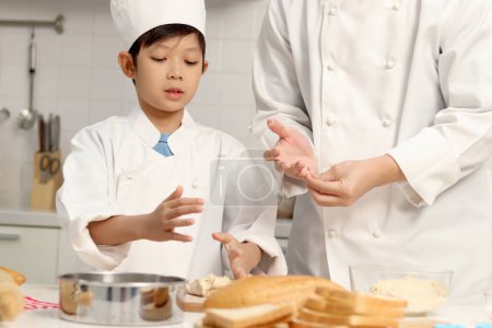 Photo for Happy Asian son and father in chef uniform with hat cook meal together at kitchen, dad parent and boy child prepare bread dough, cute family making bakery food. Kid student learns to bake a bread - Royalty Free Image