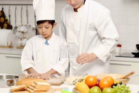 Photo for Happy Asian son and father in chef uniform with hat cook meal together at kitchen, dad parent and boy child prepare bread dough, cute family making bakery food. Kid student learns to bake a bread - Royalty Free Image