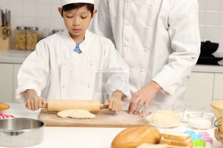 Photo for Happy Asian son and father in chef uniform with hat cook meal together at kitchen, cute boy kneads dough by rolling pin, parent and kid in family making bakery. Child student learns to bake a bread. - Royalty Free Image