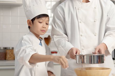 Photo for Happy Asian son and father in chef uniform with hat cooking at kitchen. cute boy child helps dad sifting flour into bowl, preparing bread dough before kneading, parent and kid in family making bakery. - Royalty Free Image