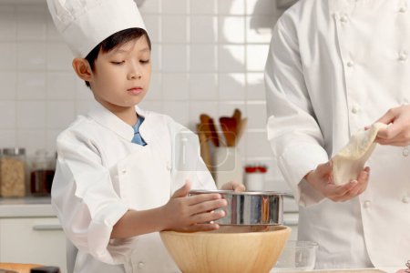 Photo for Happy Asian son and father in chef uniform with hat cooking at kitchen. cute boy child helps dad sifting flour into bowl, preparing bread dough before kneading, parent and kid in family making bakery. - Royalty Free Image