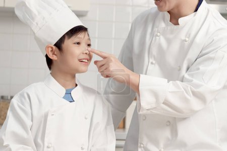 Photo for Happy Asian son and father in chef uniform with hat at kitchen, parent and boy child prepare bread dough, dad touches flour on son nose by finger, cute family have fun together during making bakery. - Royalty Free Image
