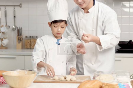 Photo for Happy Asian son and father in chef uniform with hat cook meal together at kitchen, dad parent and boy child preparing and kneading bread dough, cute family making bakery. Kid student learns to bake. - Royalty Free Image