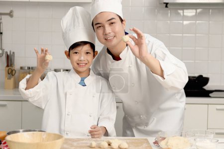 Photo for Happy Asian son and father in chef uniform with hat showing bread dough at kitchen, dad parent and boy child preparing to bake bread, cute family making bakery together. Kid student learns to bake. - Royalty Free Image