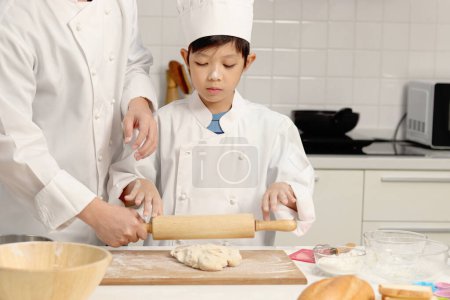 Photo for Happy Asian son and father in chef uniform with hat cook meal together at kitchen, cute boy kneads dough by rolling pin, parent and kid in family making bakery. Child student learns to bake a bread. - Royalty Free Image