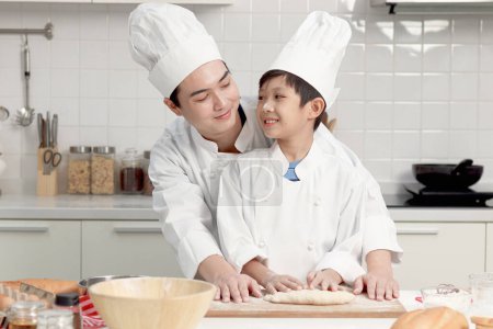 Photo for Happy Asian son and father in chef uniform with hat cooking at kitchen. Boy kneads dough by rolling pin, dad helps kid to bake a bread, look at each other during making bakery together, lovely family. - Royalty Free Image
