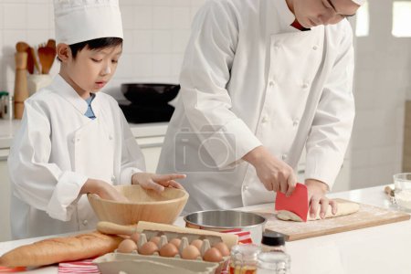 Photo for Happy Asian son and father in chef uniform with hat cook meal together at kitchen, dad parent and boy child mixing and kneading bread dough, cute family making bakery. Kid student learns to bake bread - Royalty Free Image