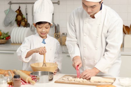 Photo for Happy Asian son and father in chef uniform with hat cook meal together at kitchen, dad parent and boy child mixing and kneading bread dough, cute family making bakery. Kid student learns to bake bread - Royalty Free Image