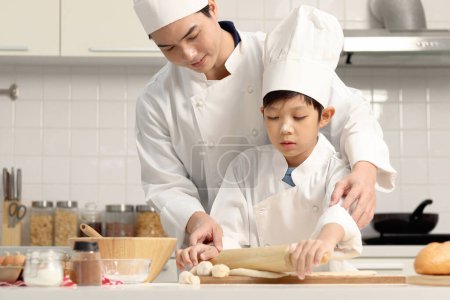 Photo for Happy Asian son and father in chef uniform with hat cook meal at kitchen, cute boy child kneads dough by rolling pin, dad helps and teaches kid to bake a bread, lovely family making bakery together. - Royalty Free Image