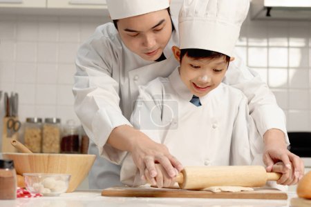 Photo for Happy Asian son and father in chef uniform with hat cook meal at kitchen, cute boy child kneads dough by rolling pin, dad helps and teaches kid to bake a bread, lovely family making bakery together. - Royalty Free Image