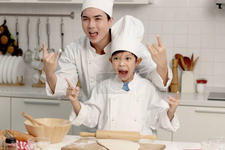 Photo for Happy Asian son kid and father in chef uniform with hat showing rock and roll finger gesture at kitchen together, dad parent and boy child having fun during baking bread, cute family making bakery. - Royalty Free Image