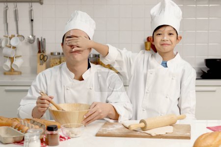 Photo for Happy Asian son kid and father in chef uniform with hat playing and having fun together at kitchen, kid covering dad eyes with his hand during baking bread, cute family making bakery. - Royalty Free Image