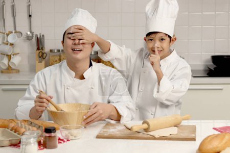 Photo for Happy Asian son kid and father in chef uniform with hat playing and having fun together at kitchen, kid covering dad eyes with his hand during baking bread, cute family making bakery. - Royalty Free Image