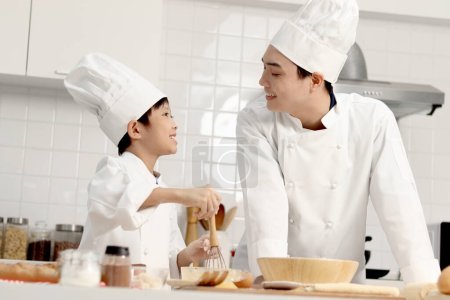 Photo for Happy Asian son and father in chef uniform with hat cook meal together at kitchen, dad parent and boy child preparing bread dough, look at each other during making bakery together, cute family. - Royalty Free Image