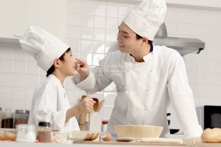 Photo for Happy Asian son and father in chef uniform with hat at kitchen, parent and boy child prepare bread dough, dad touches flour on son nose by finger, cute family have fun together during making bakery. - Royalty Free Image